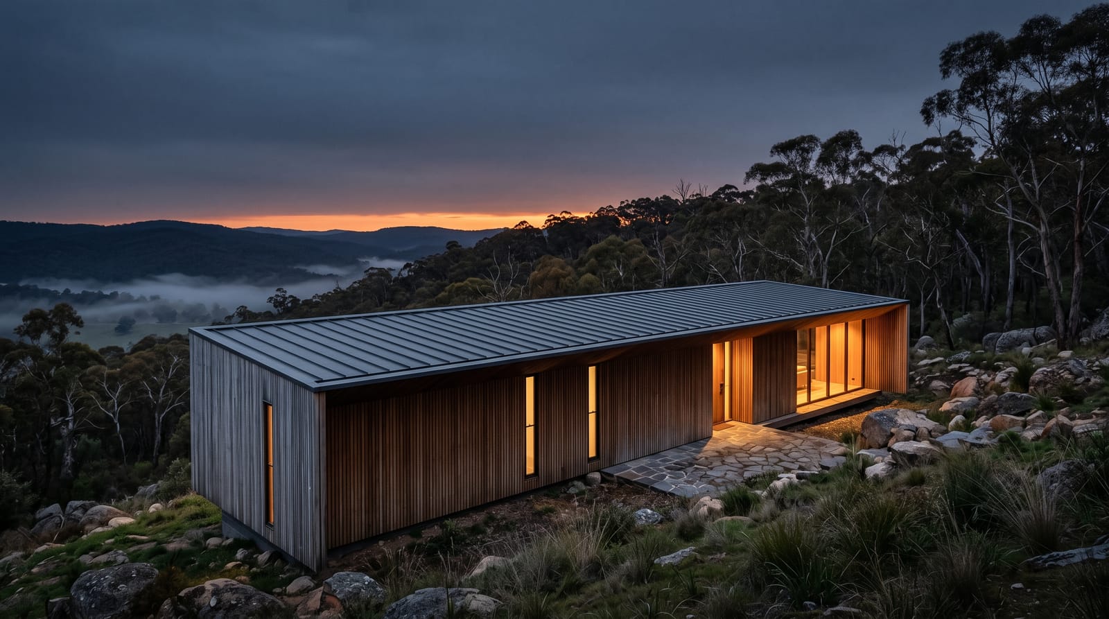 Ridgeline House at dusk: a long, low timber-clad building with a standing-seam metal roof, set among granite boulders and gum trees, its windows glowing warm above a misty valley.