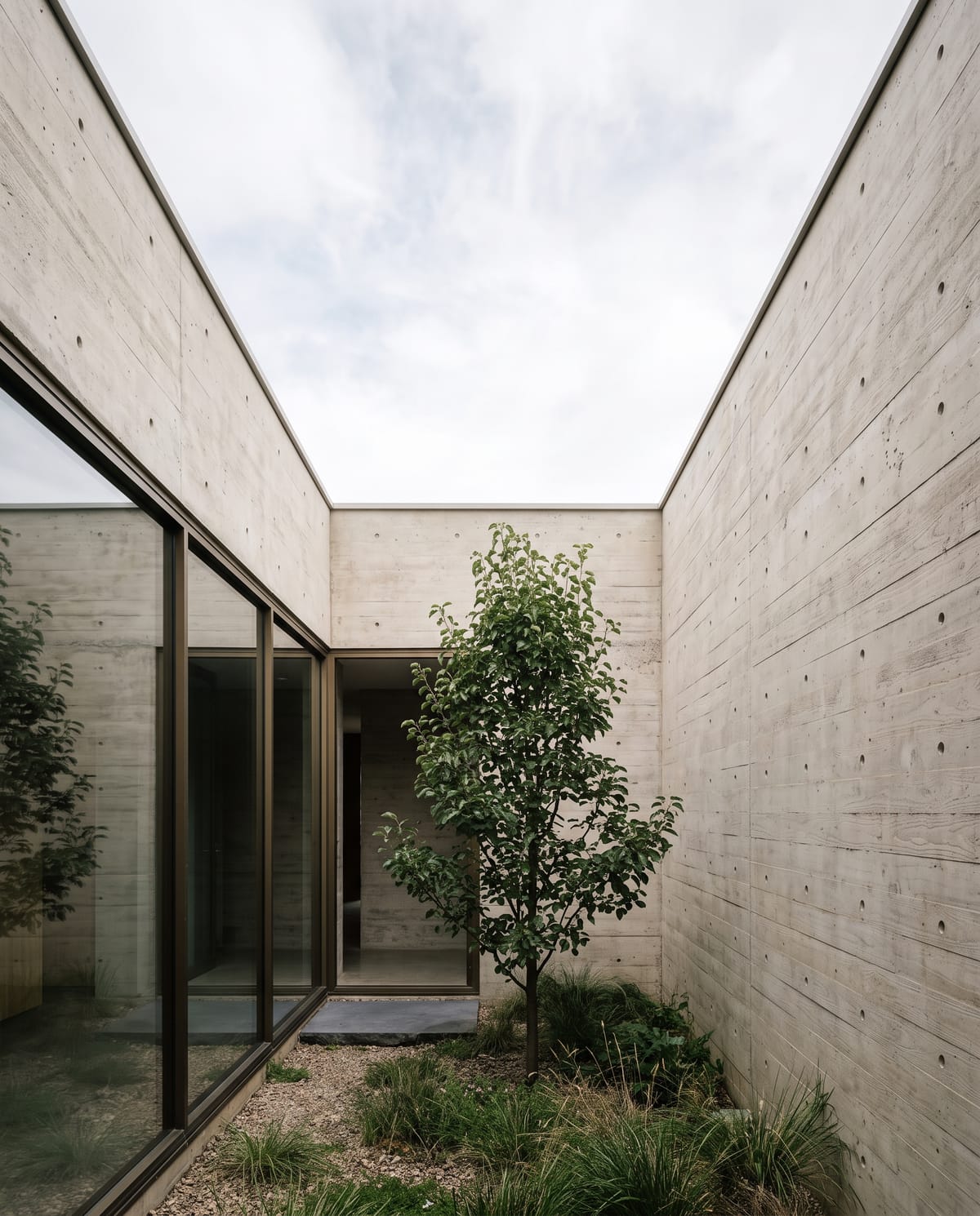 Slab House: a narrow courtyard walled in board-formed concrete, with a young tree in a gravel and grass bed beside full-height sliding glazing.